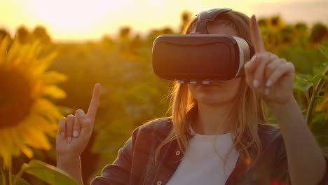 The-teenager-with-long-hair-in-plaid-shirt-and-jeans-is-working-in-VR-glasses.-She-is-engaged-in-the-working-process.-It-is-enjoyable-sunny-evening-in-the-sunflower-field-at-sunset.